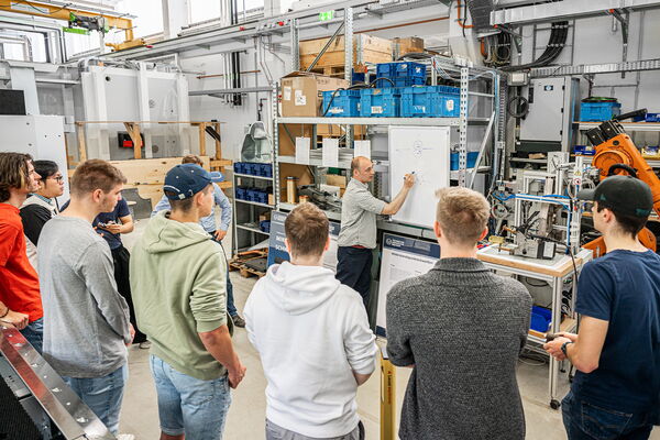 An expert is explaning something on a small board to the students standing around him, they are in a lab.