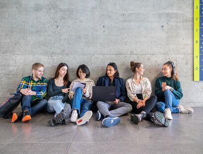 Six students are sitting on the ground and discussing inside of a campus building.