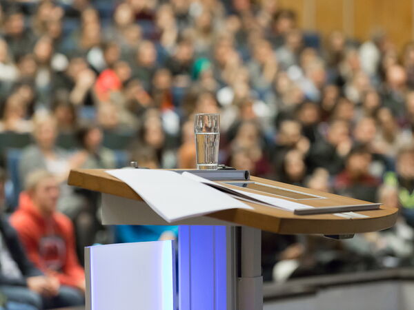 A large auditorium, in from speaker deskt with a glass of water on it and in the backgraund students are visible  with a blurry view.