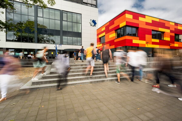 Students are walking in front of campus buldings.Two buildings are in the view. One building is colored with red, orange and yellow. The students appear  blurry because of the moevement.