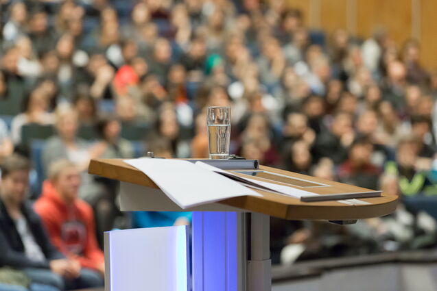A large auditorium, in from speaker deskt with a glass of water on it and in the backgraund students are visible  with a blurry view.