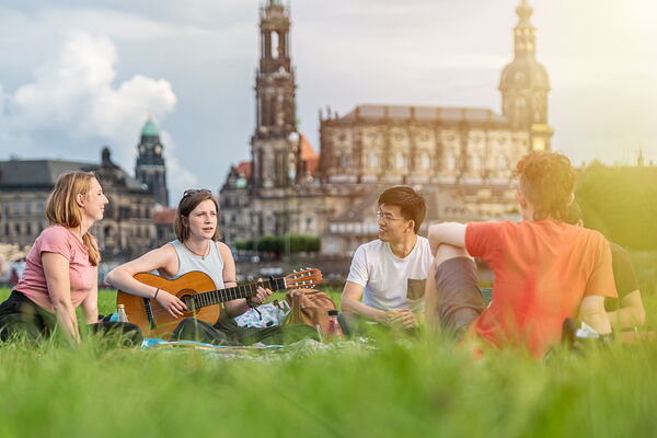 Students are sitting on grass and one of them is playing guitar and the rest is listening to her.