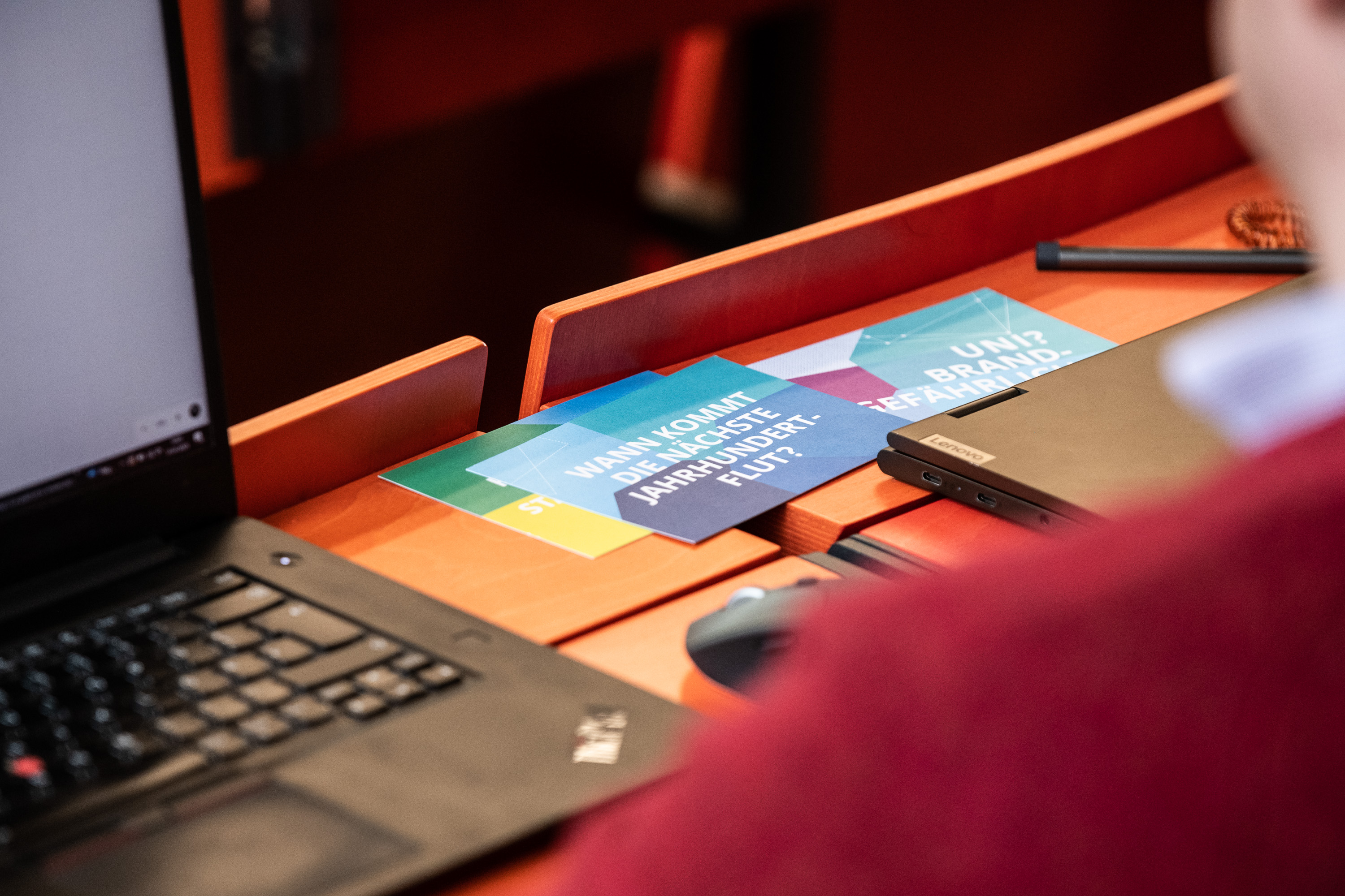 A narrow view from the shoulder of a student in a lecture room,  laptops on a desk with some brochures and front seats are in the view.