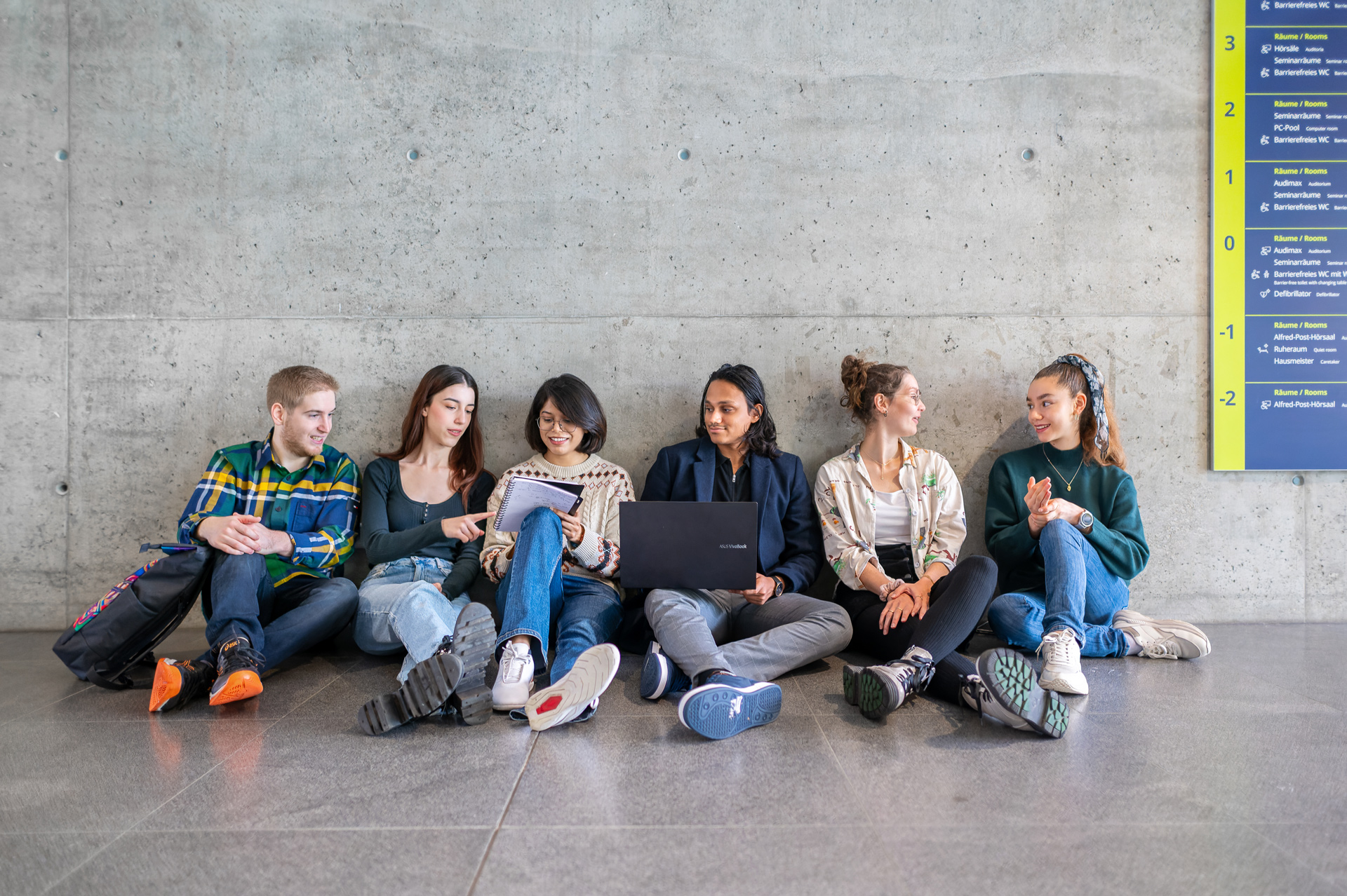 Six students are sitting on the ground and discussing inside of a campus building.